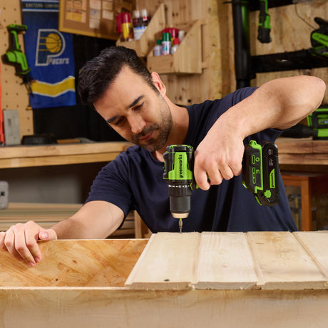 Man using a green and black cordless drill on a wooden surface in a workshop.