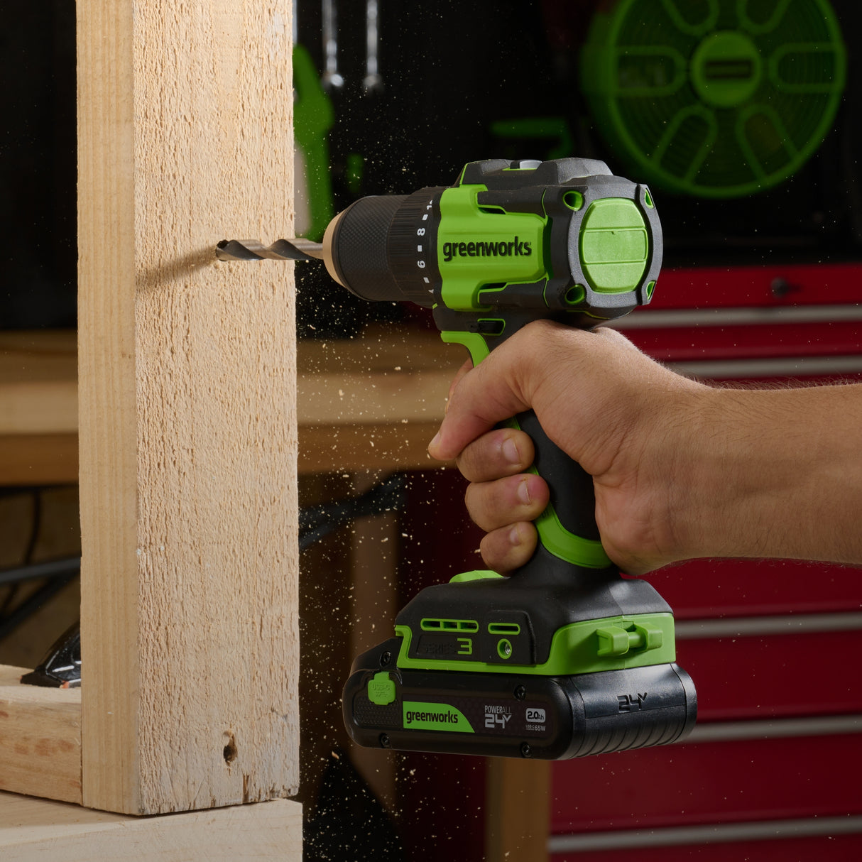 Greenworks cordless drill being used on a wooden surface with a blurred workshop background.