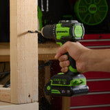 Greenworks cordless drill being used on a wooden surface with a blurred workshop background.
